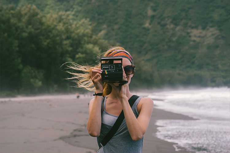 Woman in a beach holding a Polaroid camera pointing to the viewer. Say cheese!